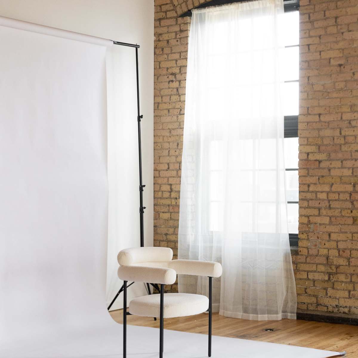 A beige chair with circular armrests is placed on a white backdrop in a bright room with large windows and exposed brick walls.