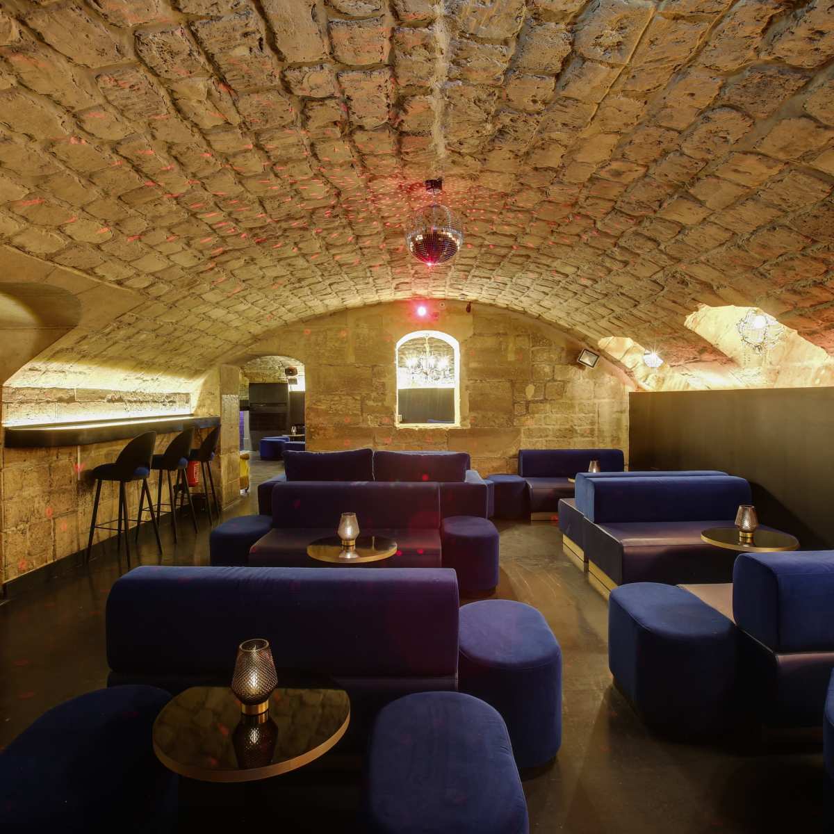 A dimly lit lounge with arched stone ceilings, blue seating arrangements, and a bar area in the background.
