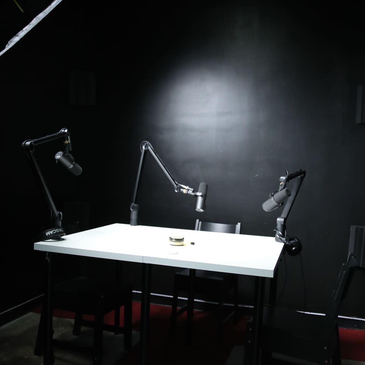 A sparse black room with a white table at the center, surrounded by four chairs and equipped with microphone stands.