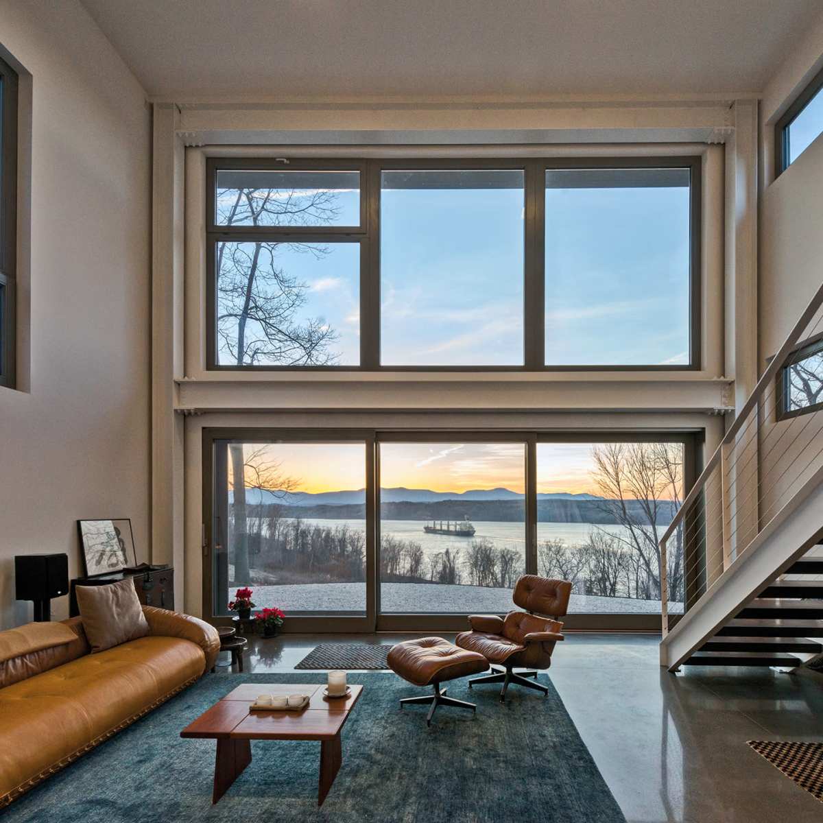 A spacious living room features large windows with a view of a lake and mountains at sunset, accompanied by a modern sofa and an armchair.