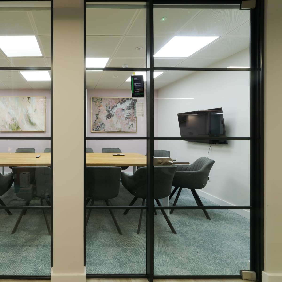 A modern conference room with a long wooden table surrounded by gray chairs is visible through glass walls, featuring two wall-mounted televisions and abstract artwork.