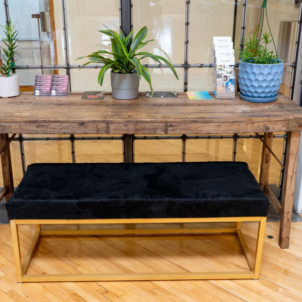 A wooden table with plants on top and a black upholstered bench with a gold frame sits in an interior space.