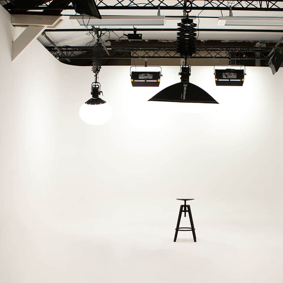 A minimalistic photography studio features a single black stool illuminated by overhead lights in a white room.