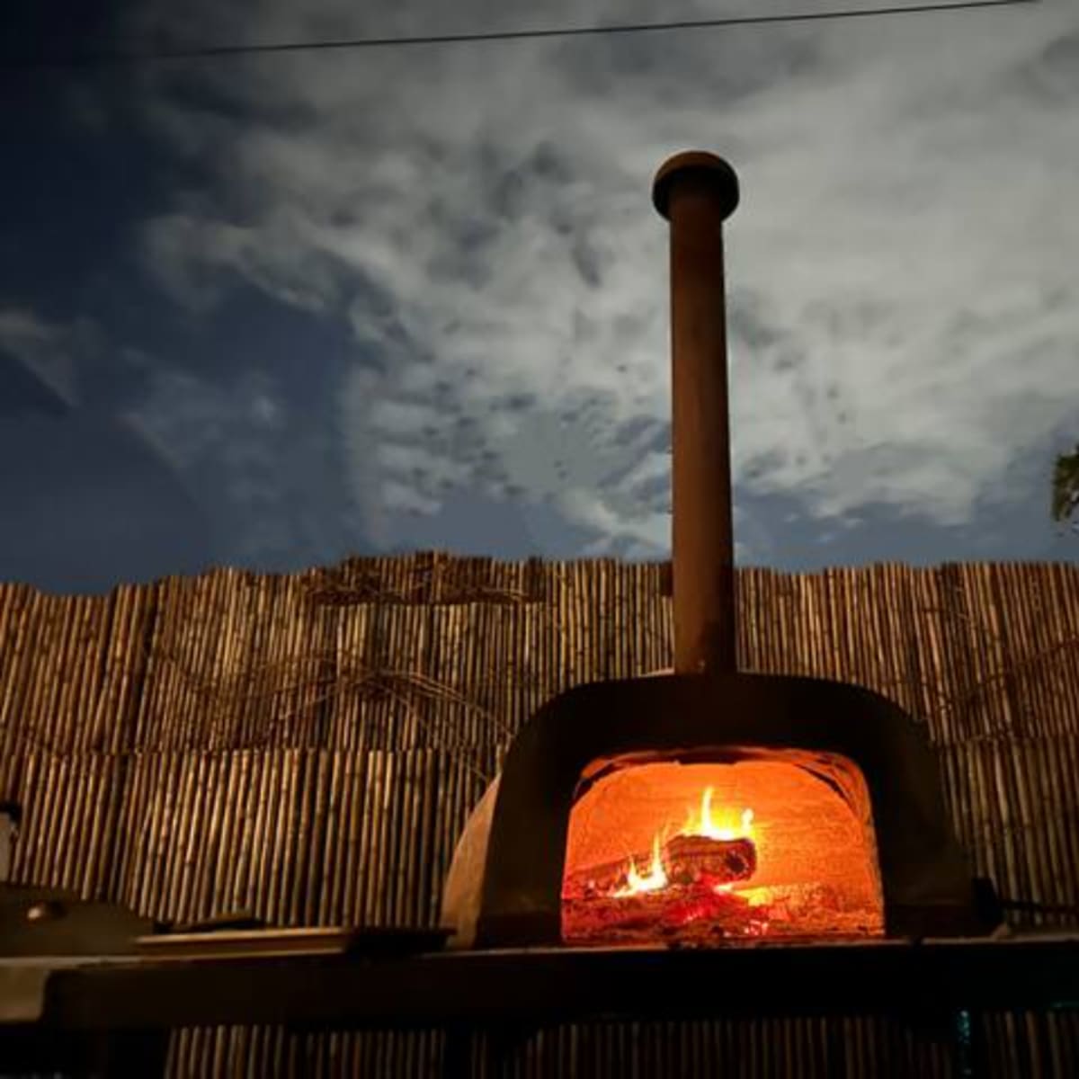 A wood-fired oven with a visible flame sits in front of a bamboo fence under a cloudy night sky.
