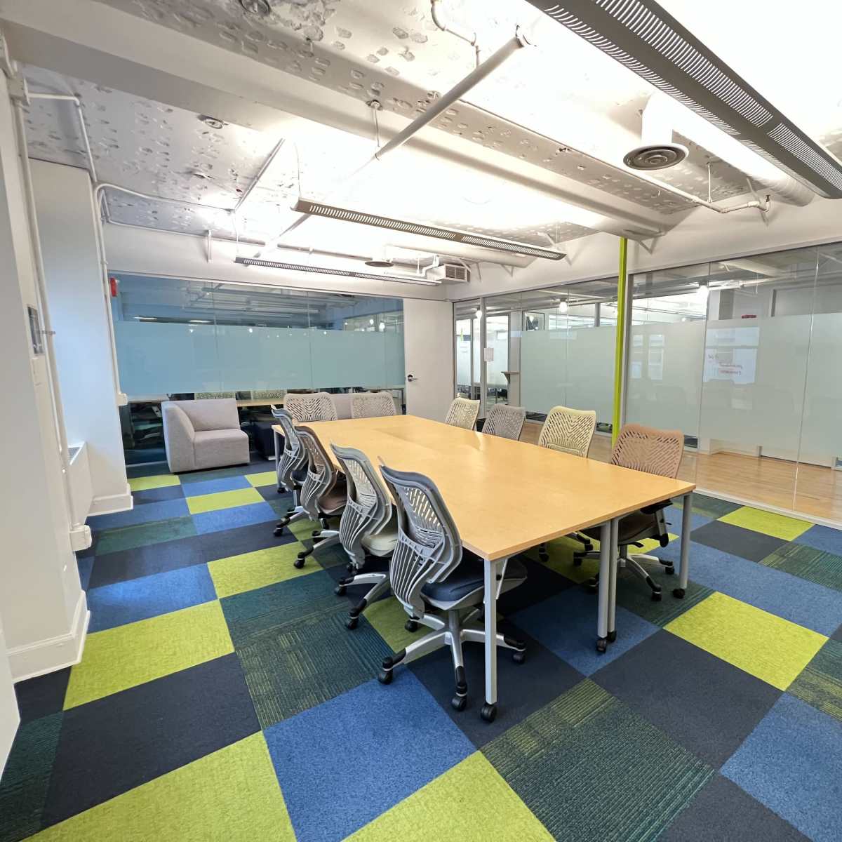 A conference room with a long wooden table surrounded by rolling chairs on a colorful carpet, adjacent to glass-paneled walls.