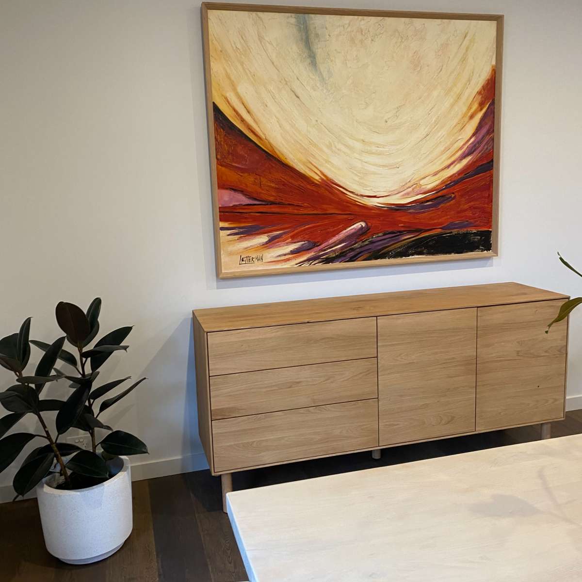 A modern interior with a wooden sideboard, a large abstract painting on the wall, and a potted plant in a white planter.