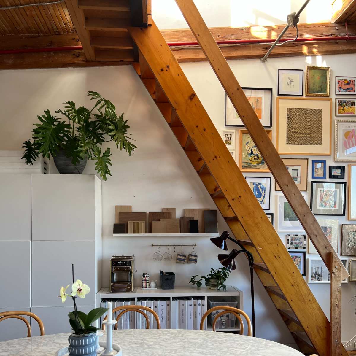 A dining area features a round marble table surrounded by bentwood chairs, with a wooden beam structure and a wall adorned with various framed artworks.