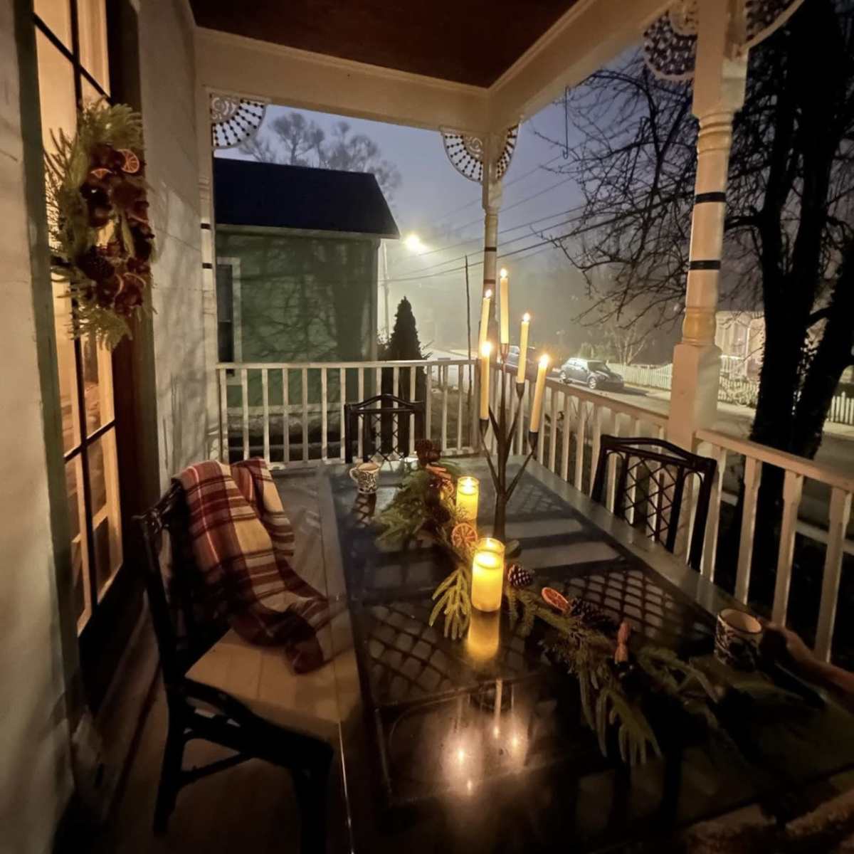The porch is decorated with a green wreath and features a table set for dining with candles and a winter floral arrangement, illuminated by soft light in a foggy atmosphere.