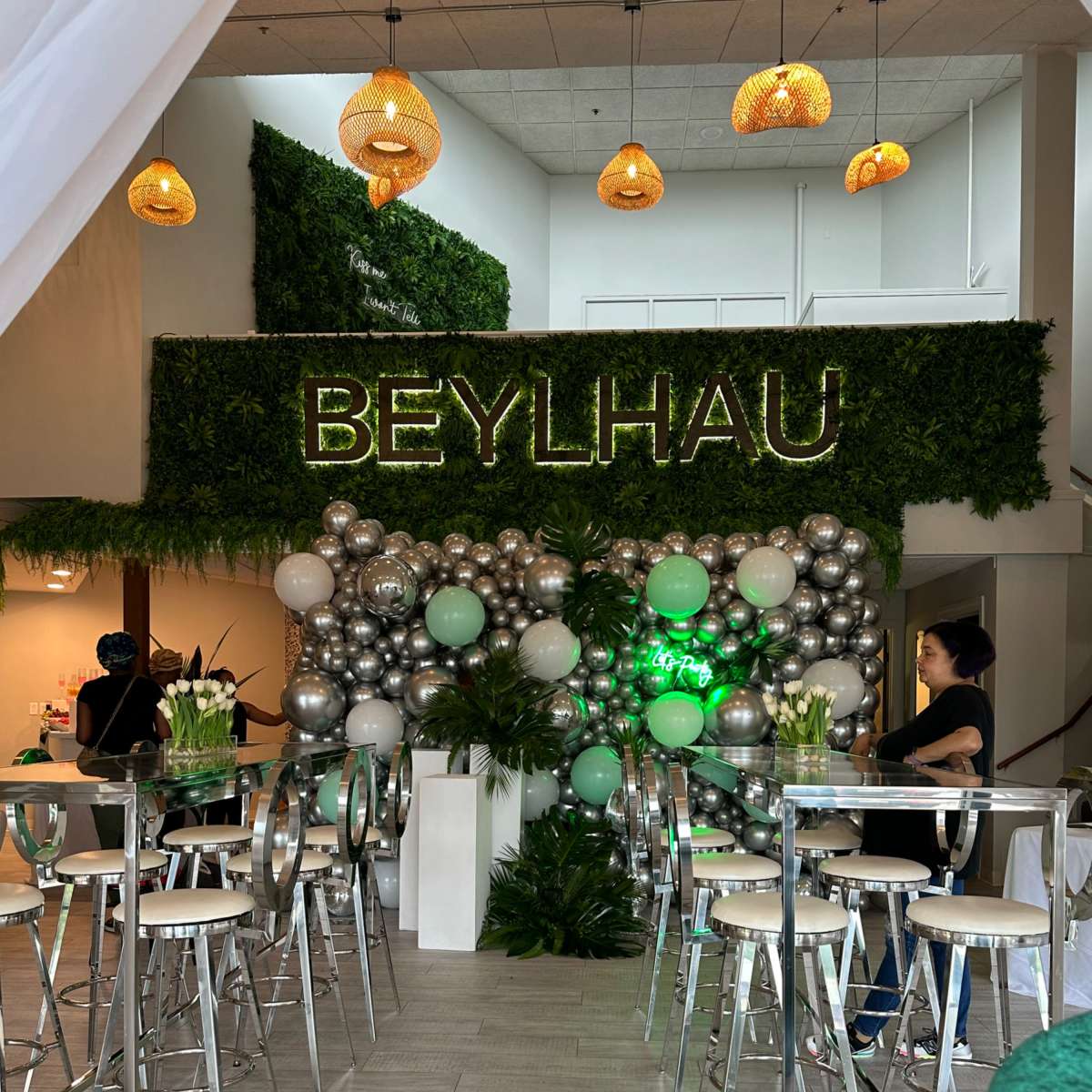An event space decorated with a greenery wall and a balloon arrangement, featuring a sign that reads "BEYLHAU," along with transparent bar stools and tables.
