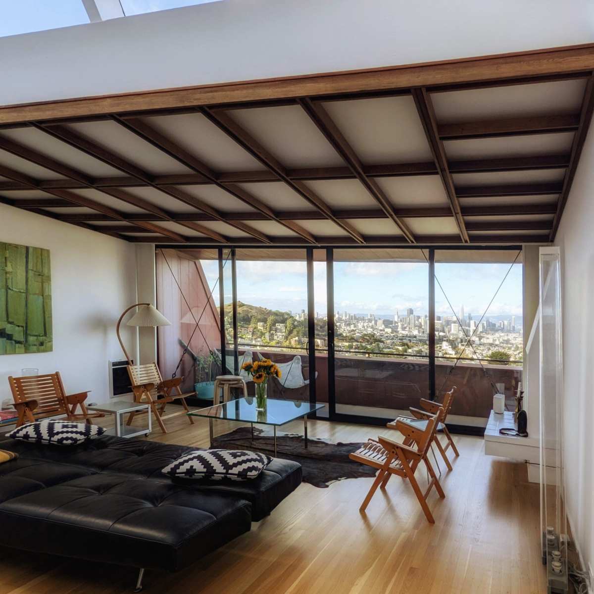 A modern living room with large glass windows overlooking a cityscape, featuring contemporary furniture and wooden ceiling beams.