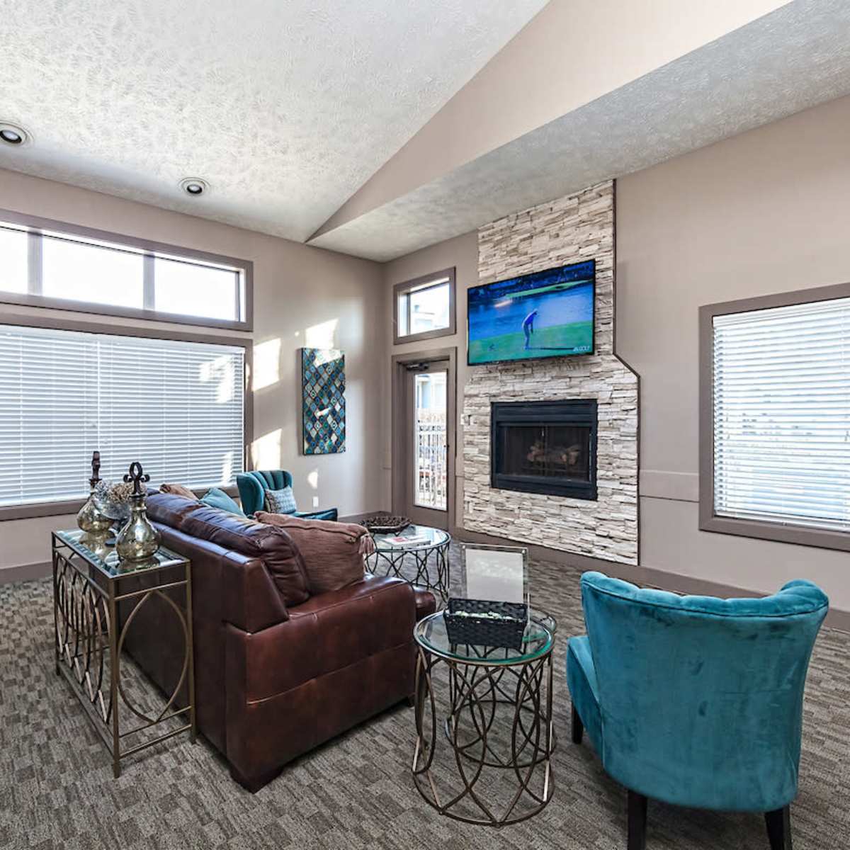 A modern living room with a brown leather sofa, a stone fireplace, and large windows allowing natural light to enter the space.