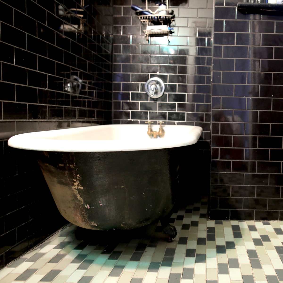 A bathroom featuring a vintage clawfoot bathtub and a modern toilet, with black tiled walls and a mosaic tile floor.