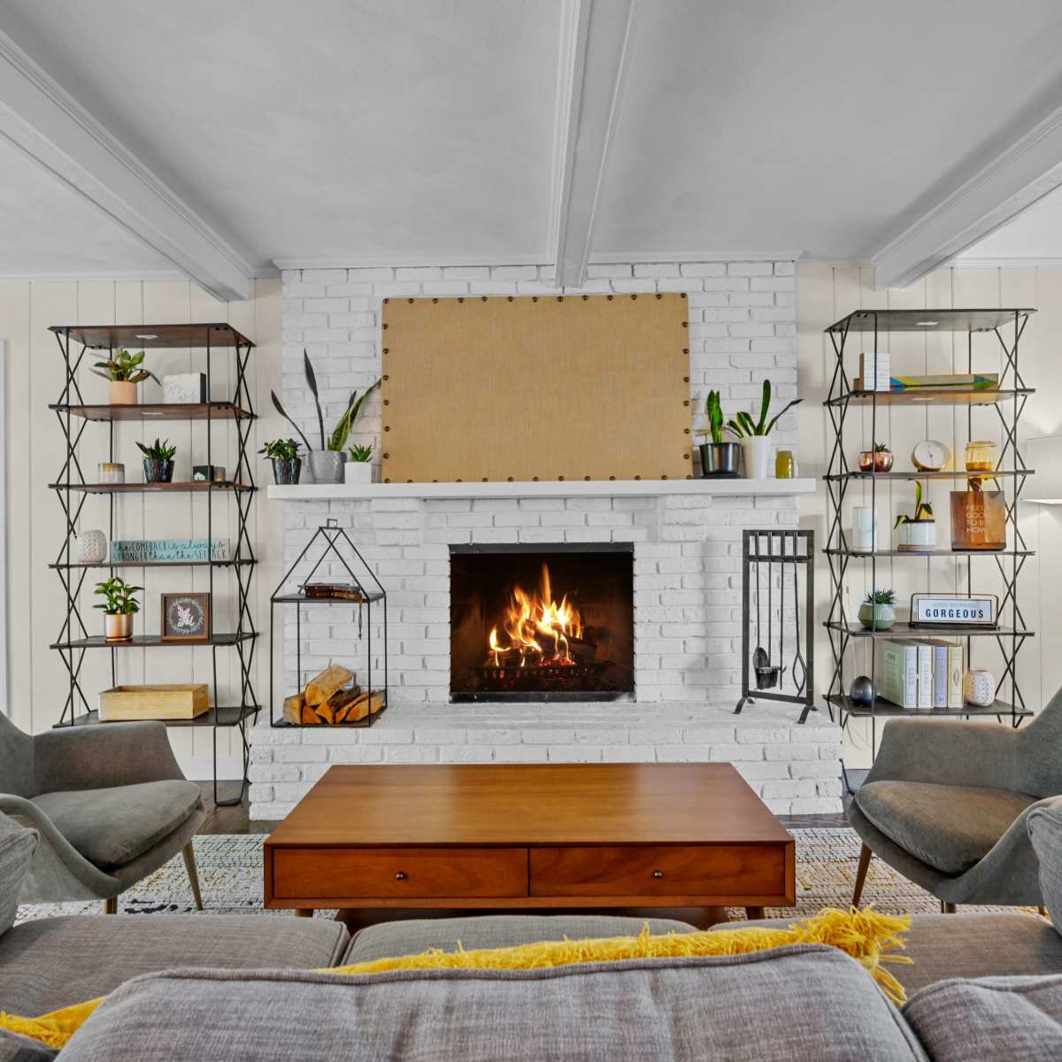 A cozy living room features a white brick fireplace surrounded by shelving units, a wooden coffee table, and two gray armchairs, with a lit fire in the hearth.