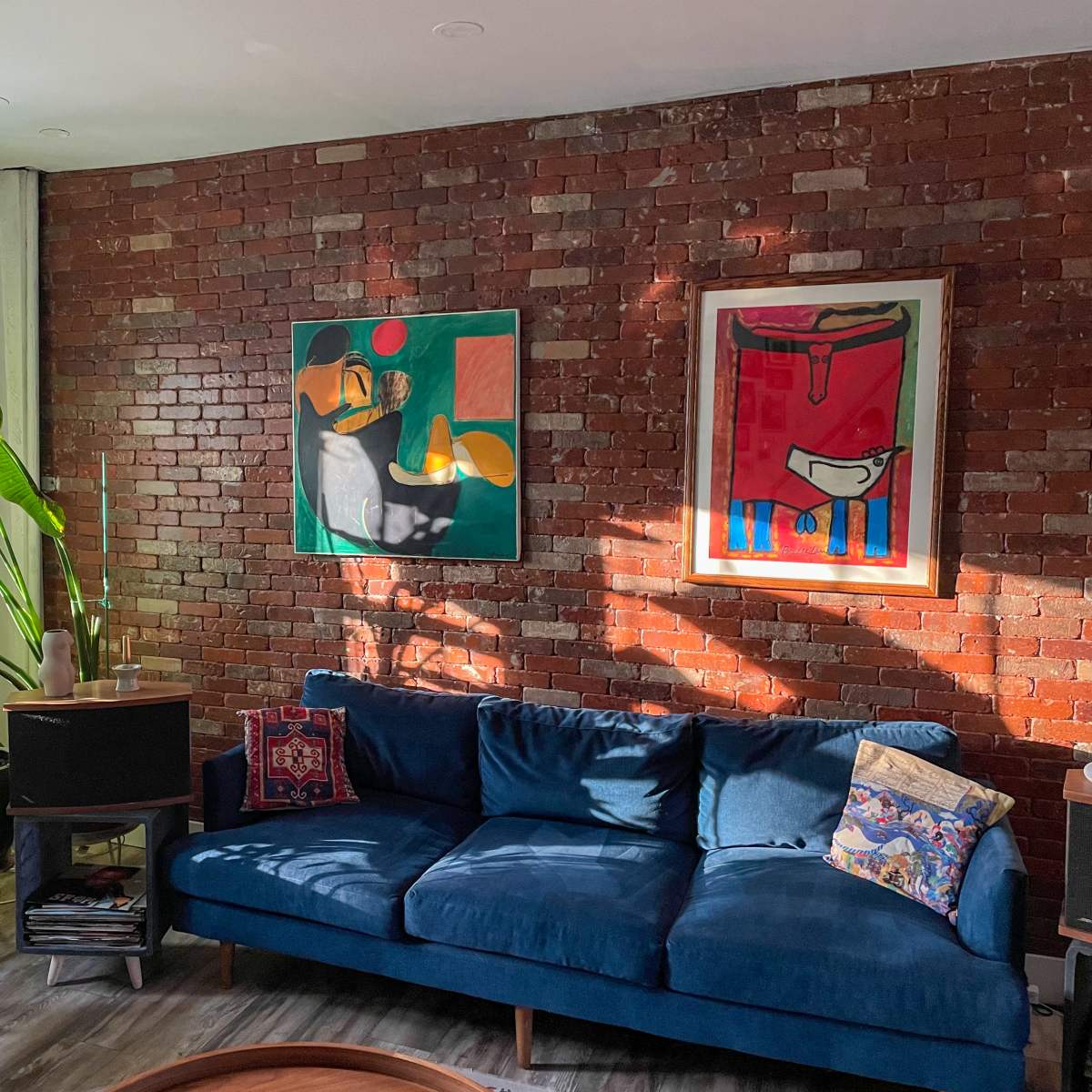 A blue velvet sofa is positioned against a brick wall, adorned with two colorful abstract paintings and surrounded by various indoor plants.