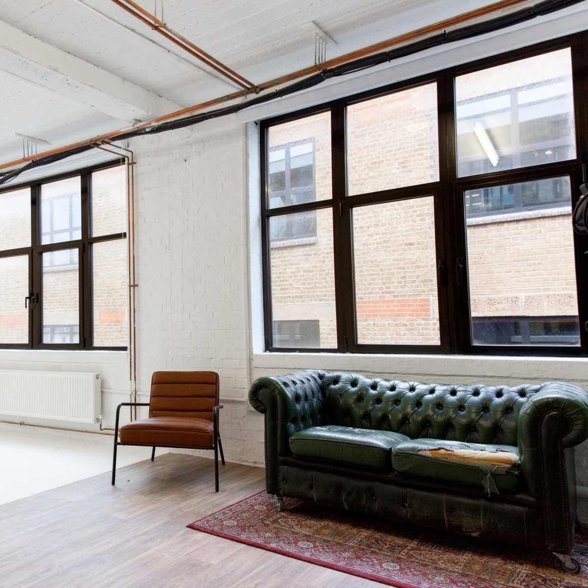 A spacious room with large windows, a green leather sofa, a brown chair, and a mirror against a white wall.
