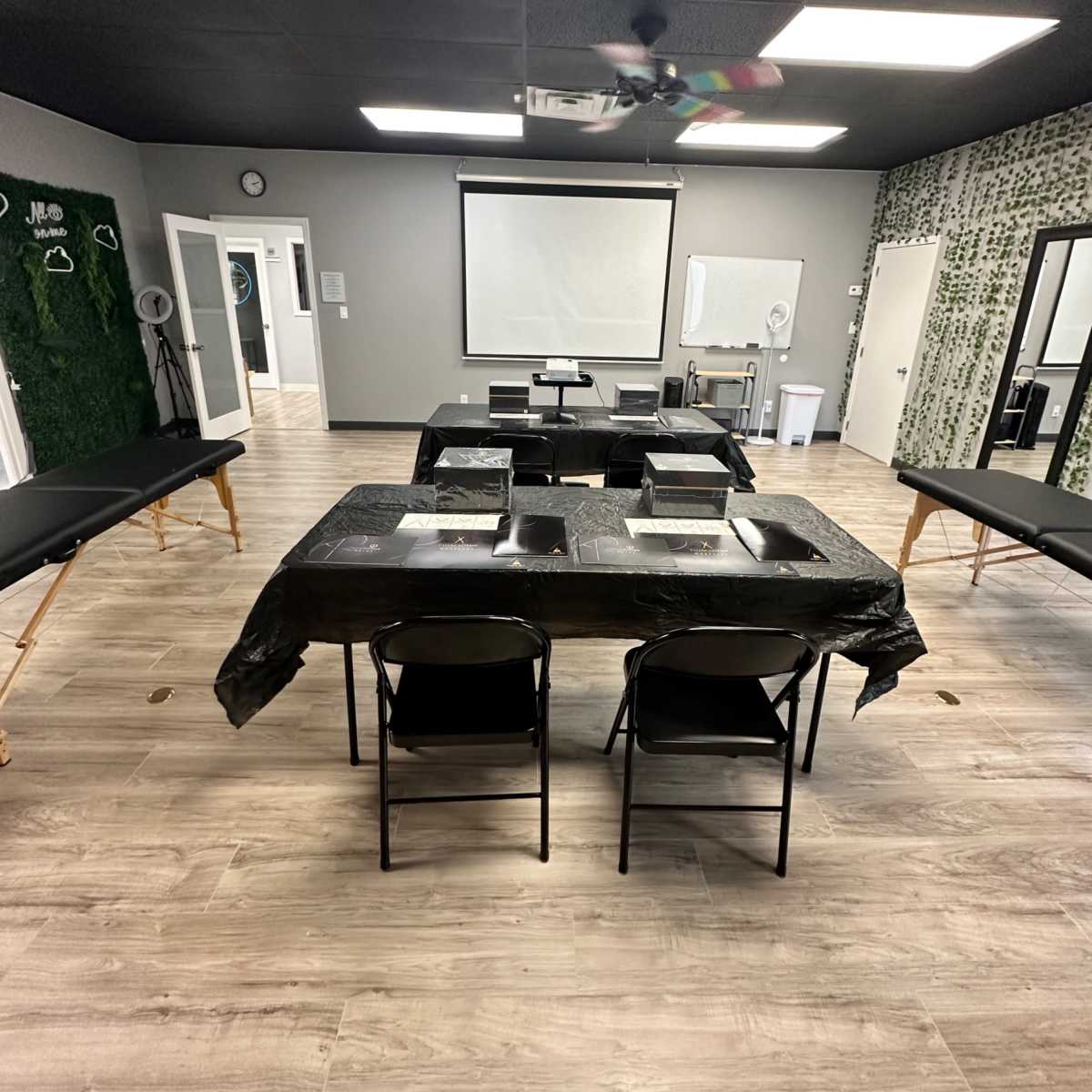 A training room set up with a central table covered in black cloth, surrounded by black folding chairs, and equipped with a projector and screen at one end.
