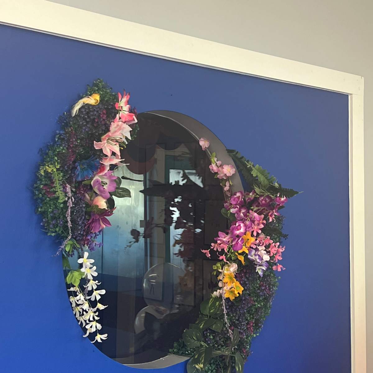 A round mirror surrounded by a floral wreath is mounted on a blue wall beneath a silhouette of a dancer.