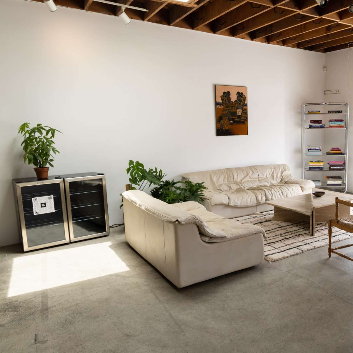 A minimalist living room with a white leather sofa, a wooden coffee table, a plant in the corner, a small shelving unit with books, and a door leading outside.