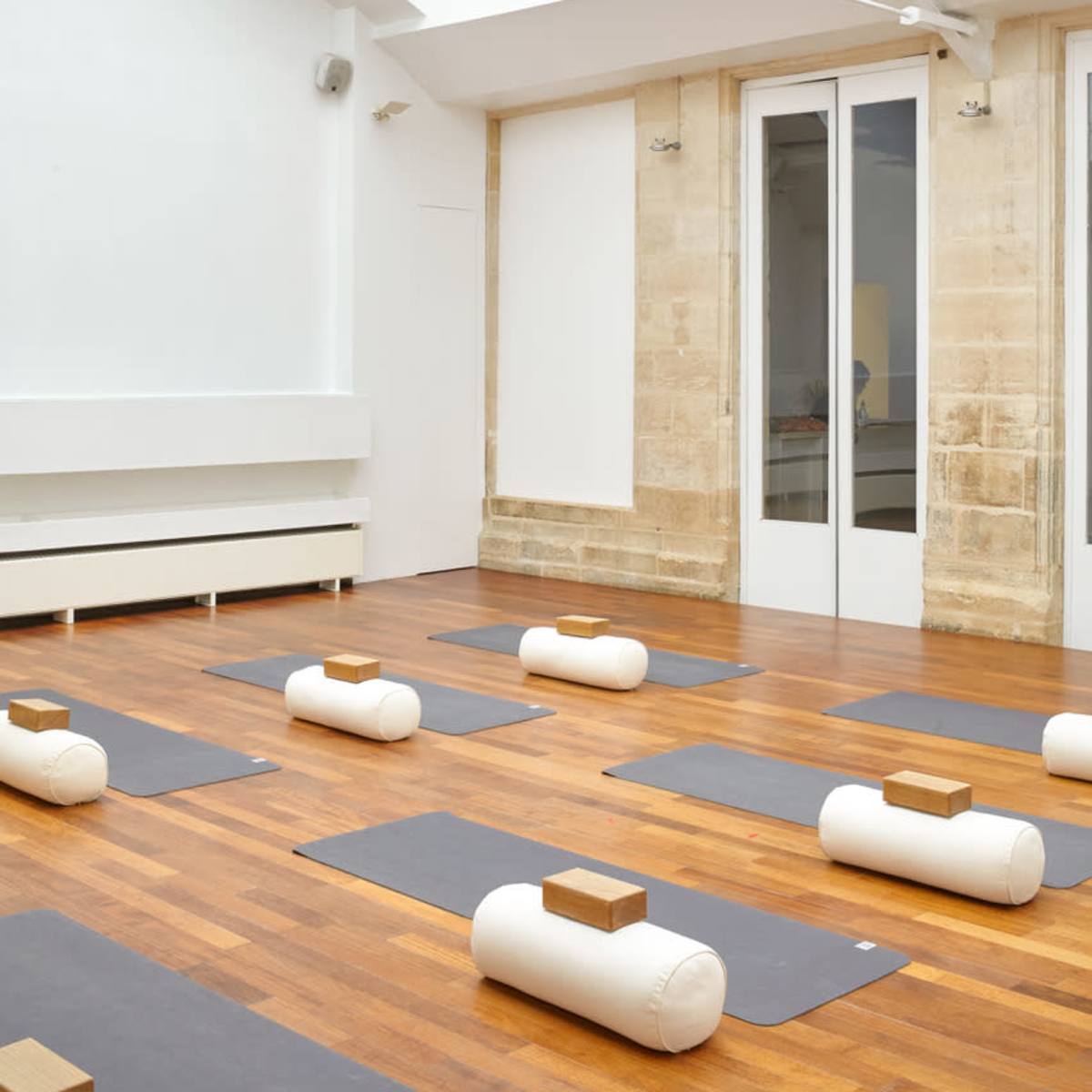 A minimalistic yoga studio with wooden floors, several yoga mats arranged on the ground, and large windows letting in natural light.