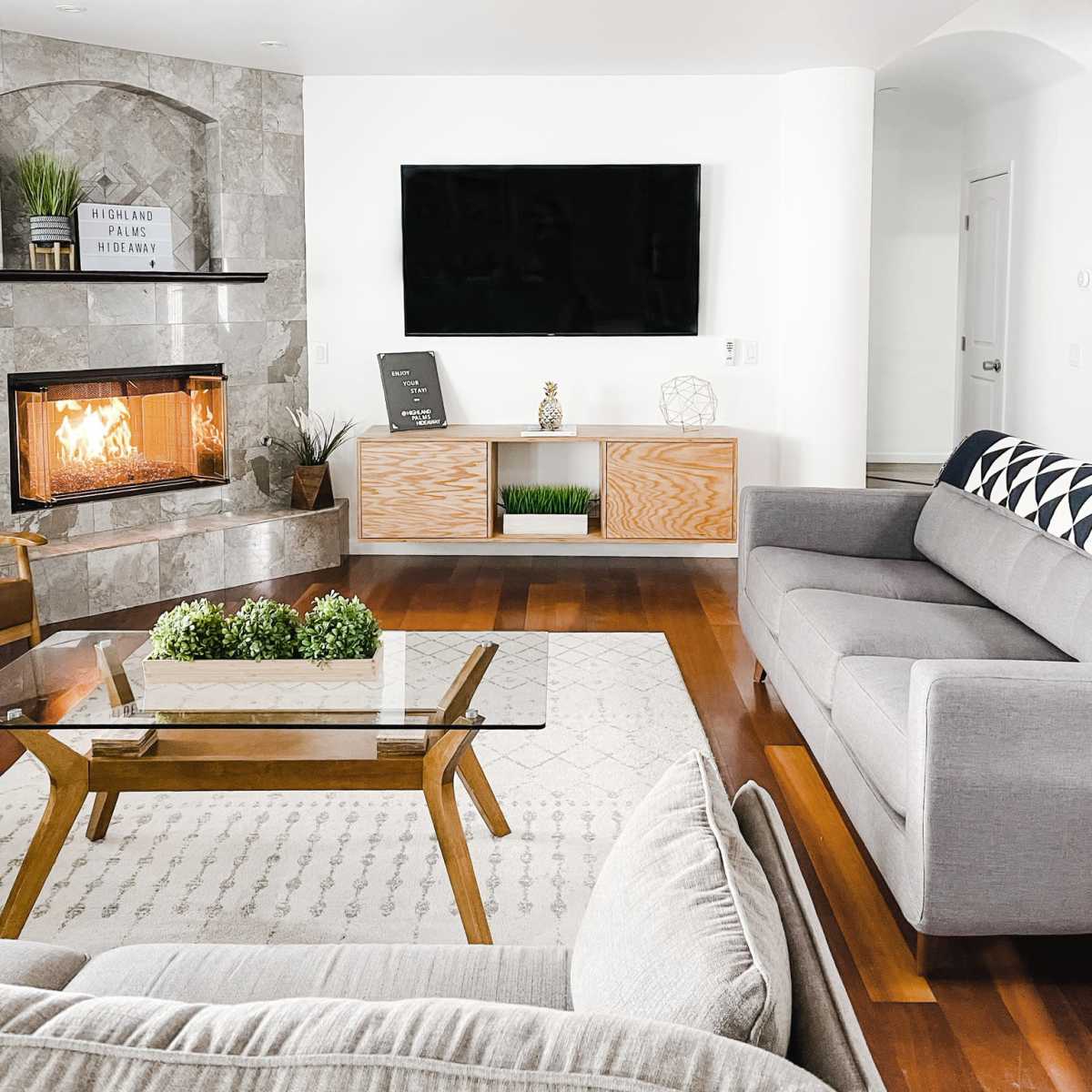 The living room features a modern design with a stone fireplace, a large television mounted on the wall, and various furniture pieces including a gray sofa and a wooden coffee table.