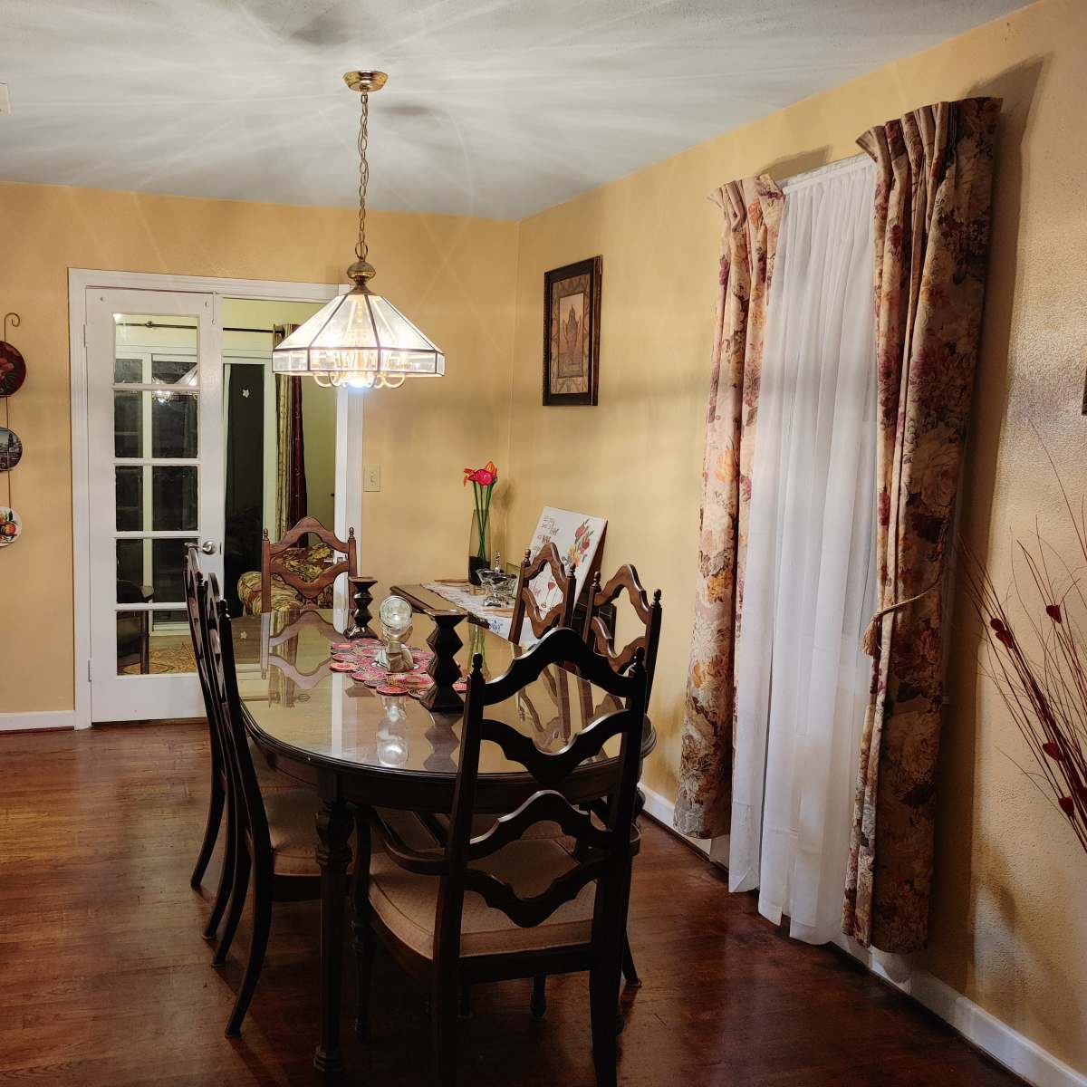 A dining room features a circular glass table surrounded by wooden chairs, illuminated by a pendant light, with a doorway leading to another room.