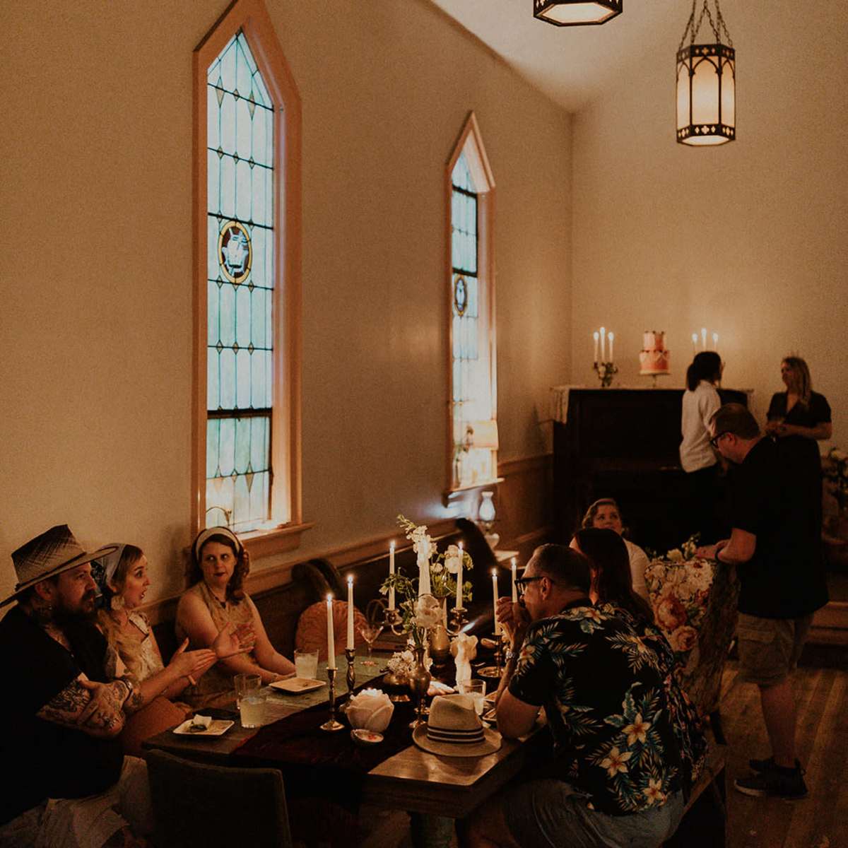 A group of people sits around a table in a dimly lit room with stained glass windows and candlelight.