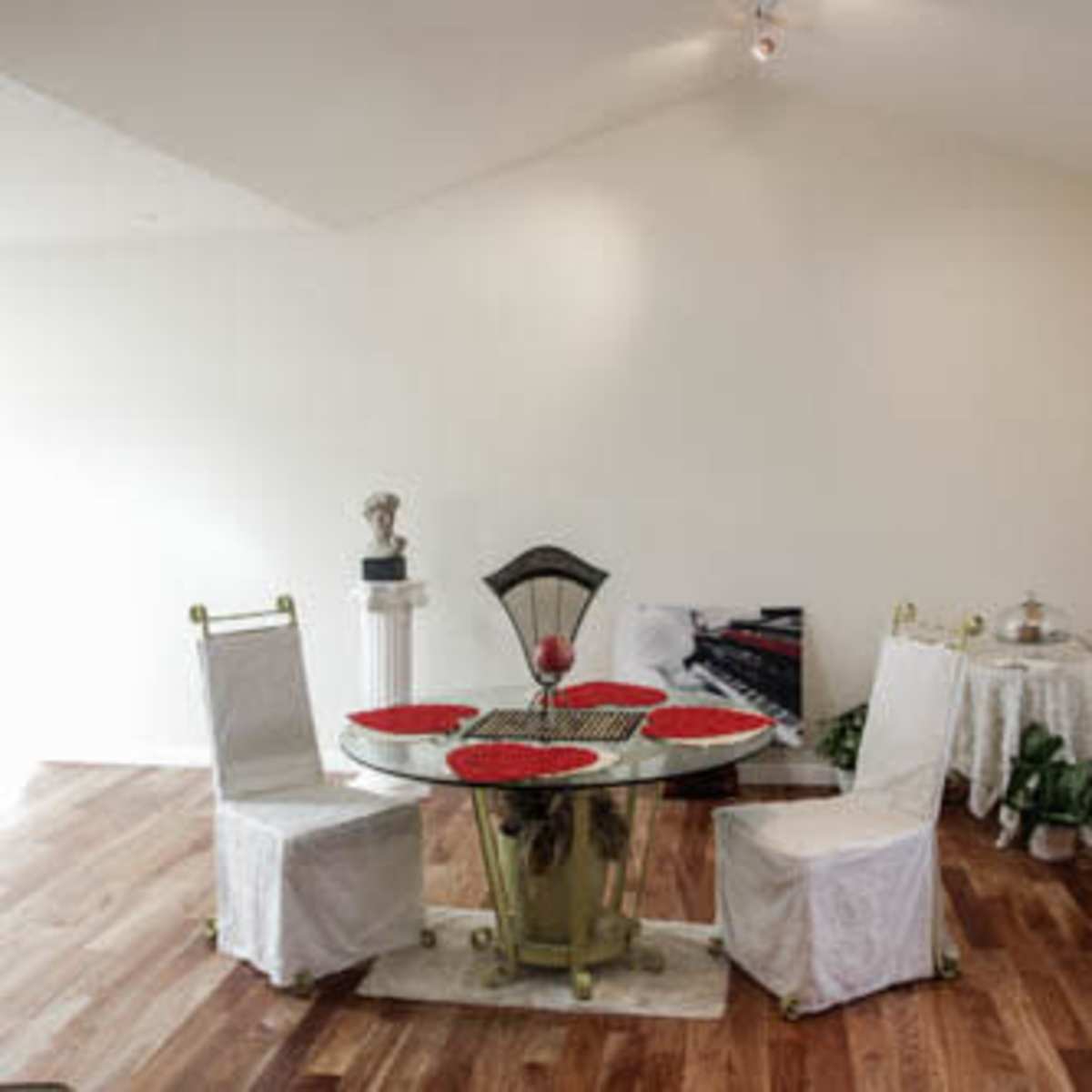 A dining area with a round table set for four, featuring red placemats, and surrounded by white-covered chairs, with a light-colored wooden floor and minimal decor.