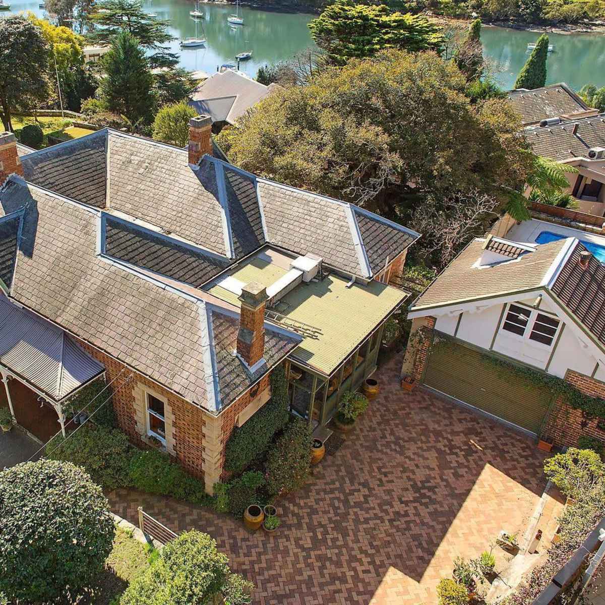A large, two-story brick house with multiple gables, surrounded by greenery and a view of a body of water in the background.