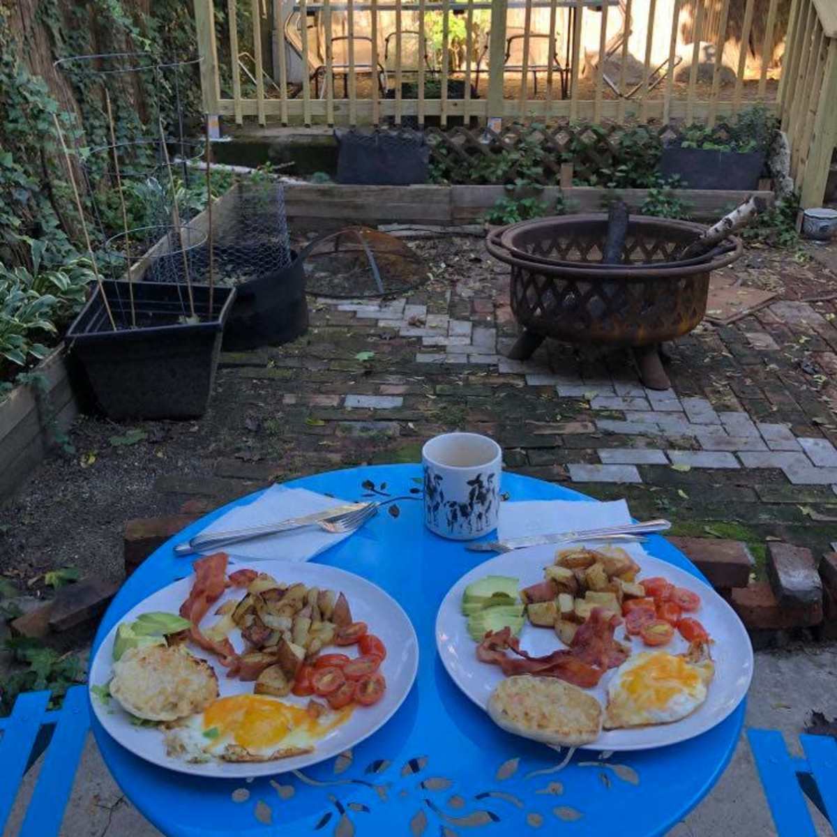 Two plates of breakfast with eggs, bacon, and potatoes are set on a blue table in a garden, with a fire pit and patio area visible in the background.