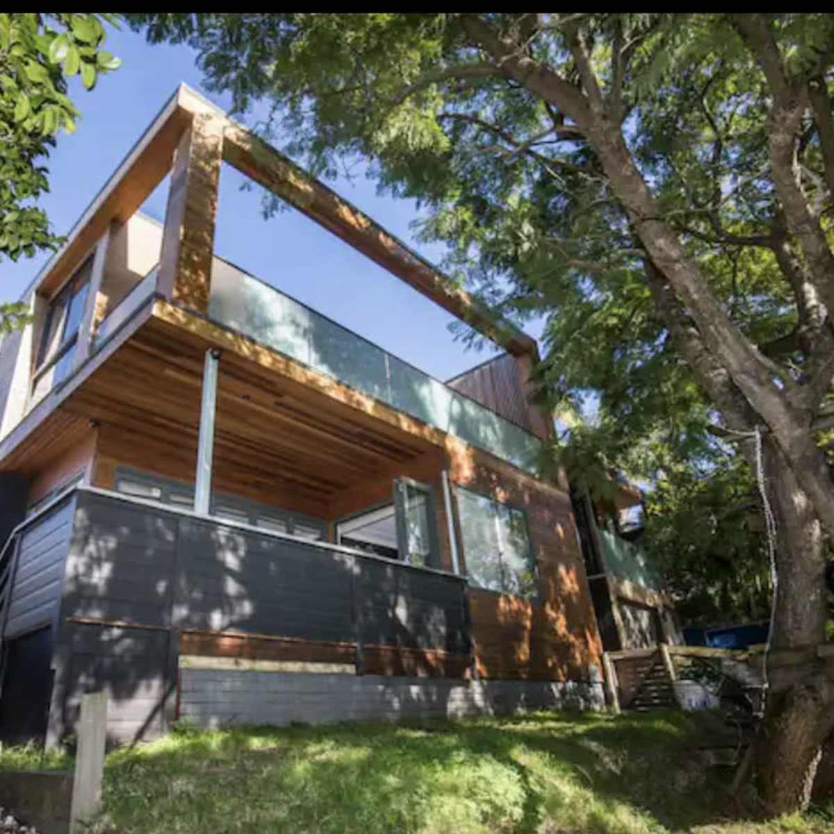 A modern two-story house with a wooden exterior, large windows, and a balcony, situated among trees and grass.