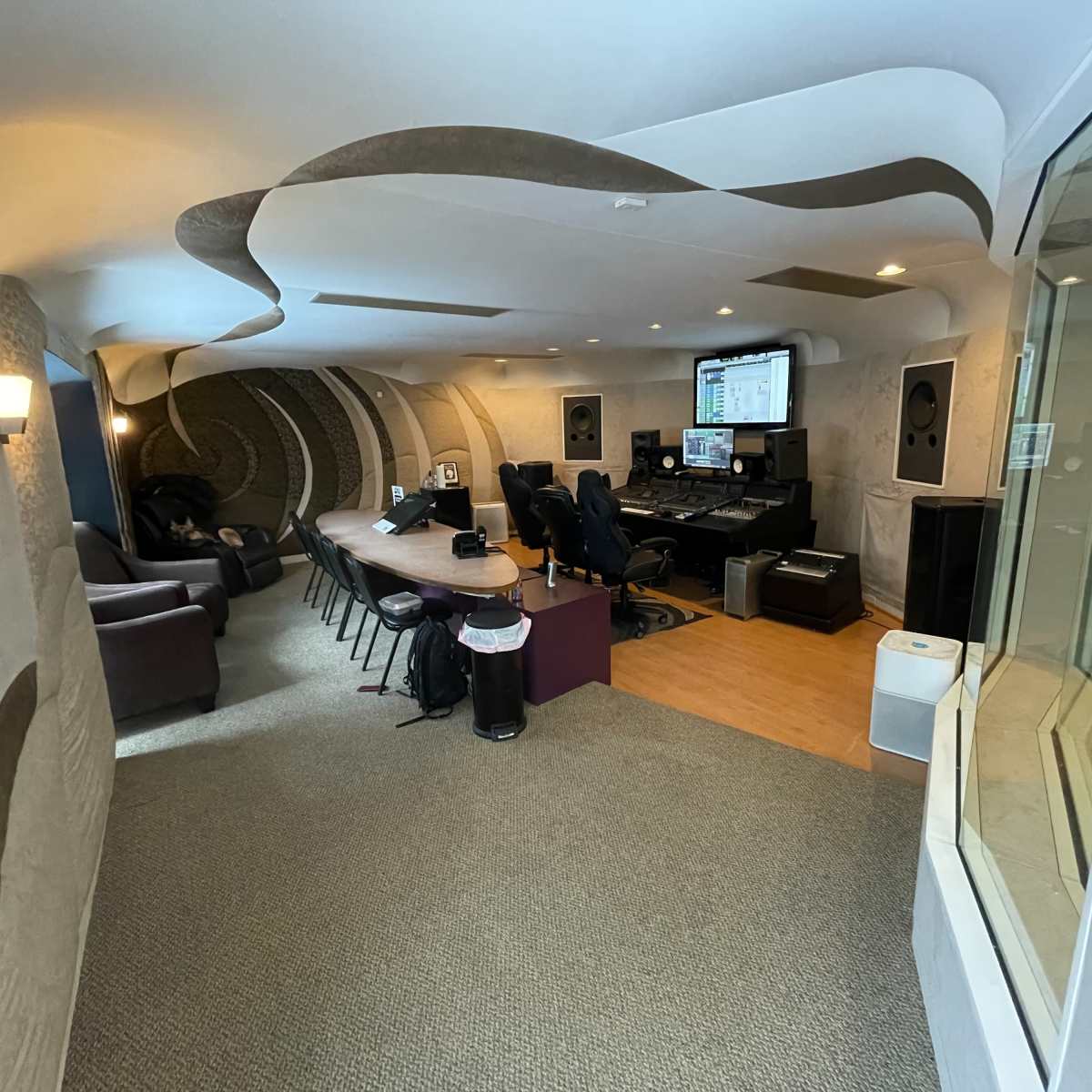 A modern recording studio featuring an elongated room with a sound mixing area, seating, and glass-walled isolation space.
