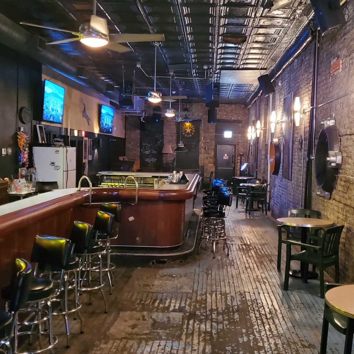 An empty bar with a wooden counter, metal stools, and exposed brick walls, illuminated by dim lighting and featuring a few screens on the walls.