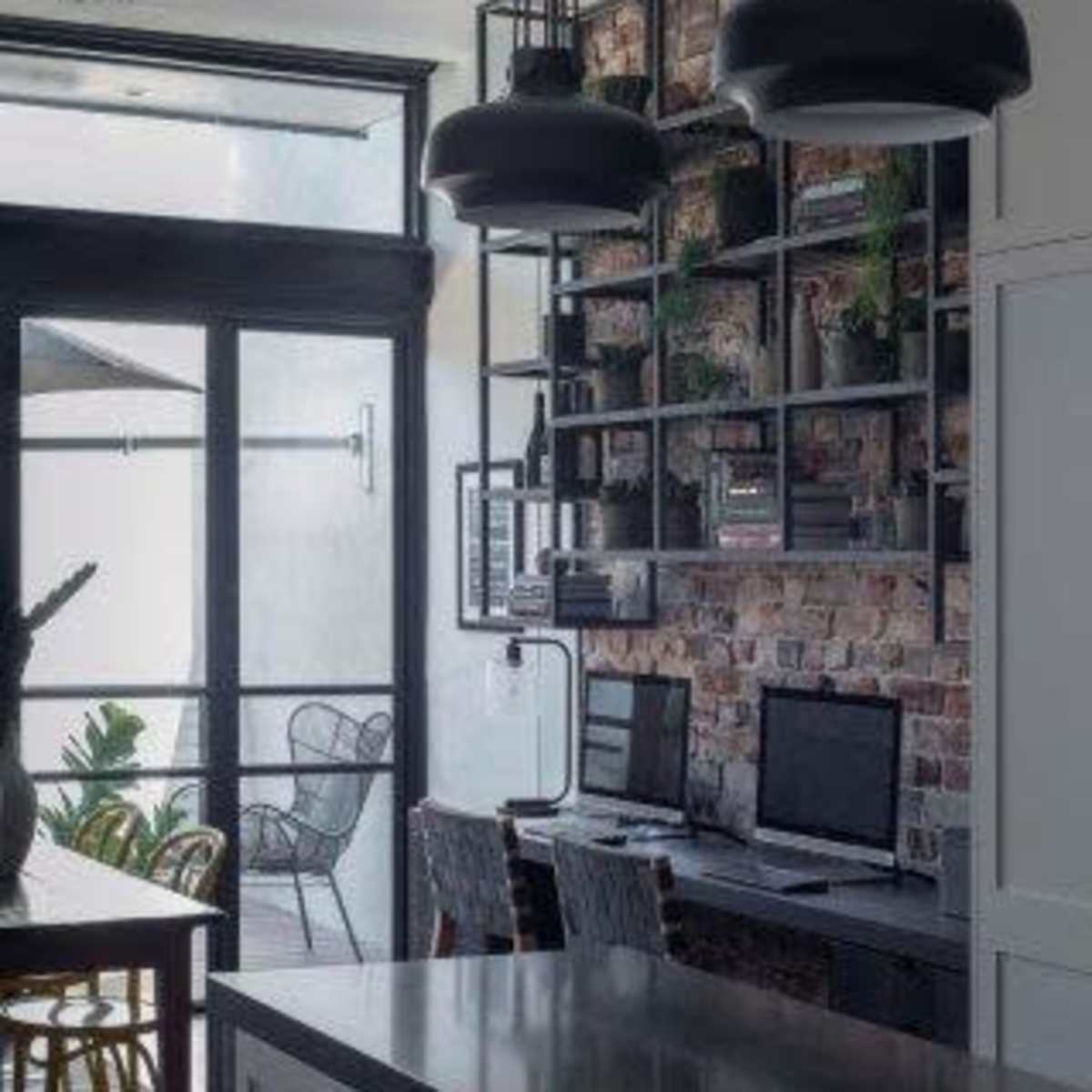 A contemporary kitchen with a dark-toned countertop, dual computers on a desk against a brick wall, and hanging lights above.