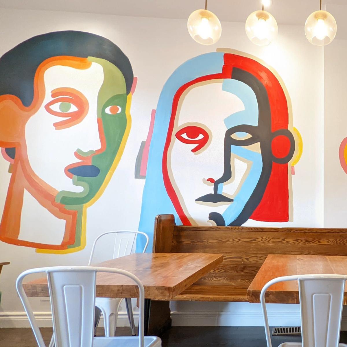 A café interior with wooden tables and chairs, illuminated by pendant lights, and a wall adorned with colorful abstract portraits.