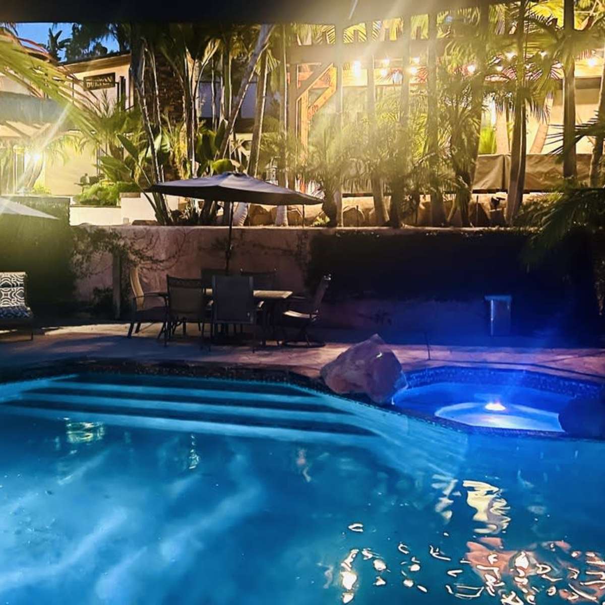 A serene backyard pool area features a lit pool, lounge chairs, and a patio surrounded by tropical plants and trees at dusk.