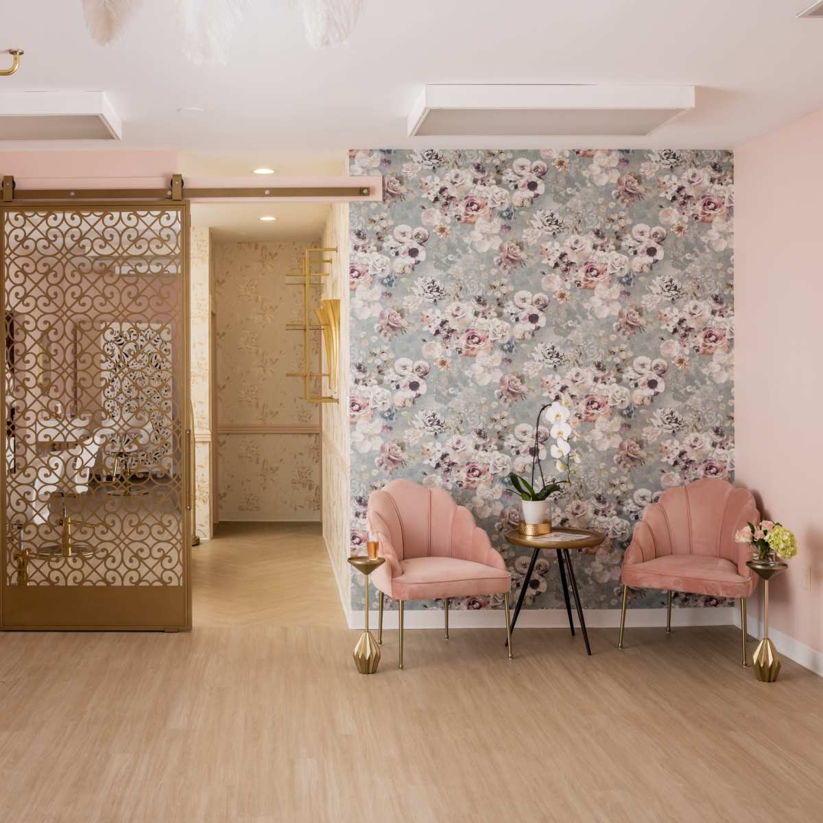 A stylish salon interior featuring gold accents, pink chairs, a decorative partition, and floral wallpaper.