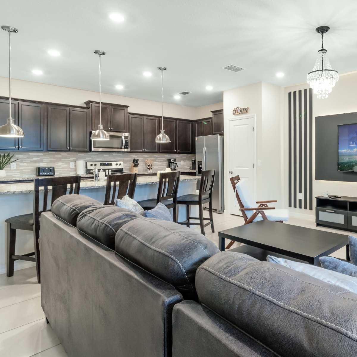 A modern living room features a sectional sofa, a coffee table, and a wall-mounted television, with a kitchen area visible in the background.
