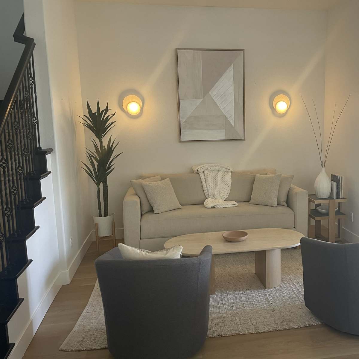 A light-filled living room features a gray sofa, two matching chairs, a round coffee table, and wall-mounted lights against a neutral backdrop.
