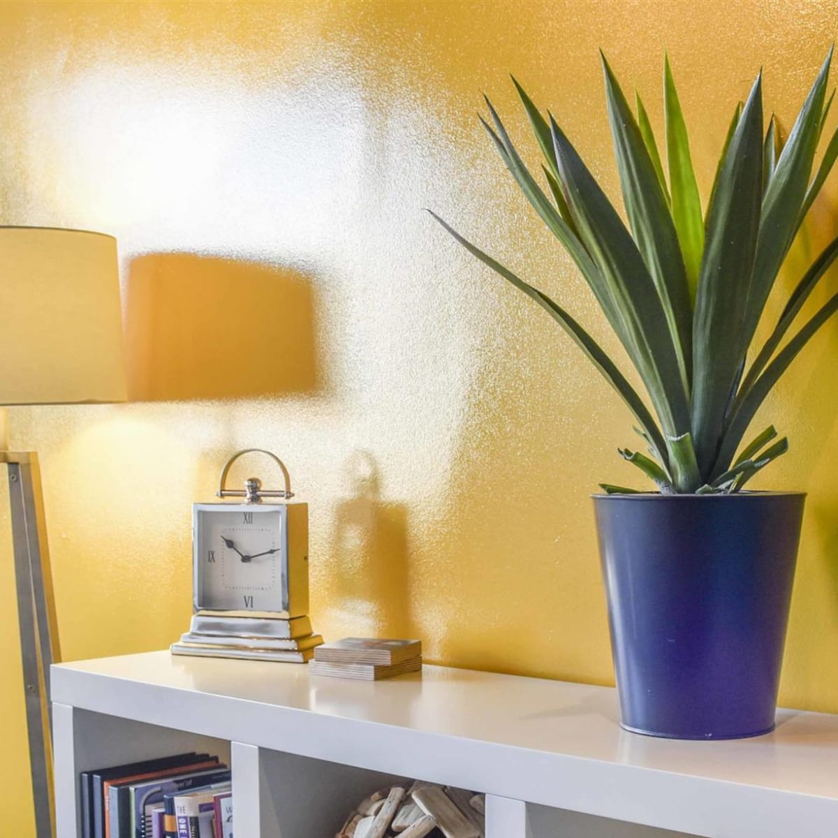 A tall plant is in a dark blue pot on a white shelf against a shiny yellow wall, with a lamp and a clock nearby.