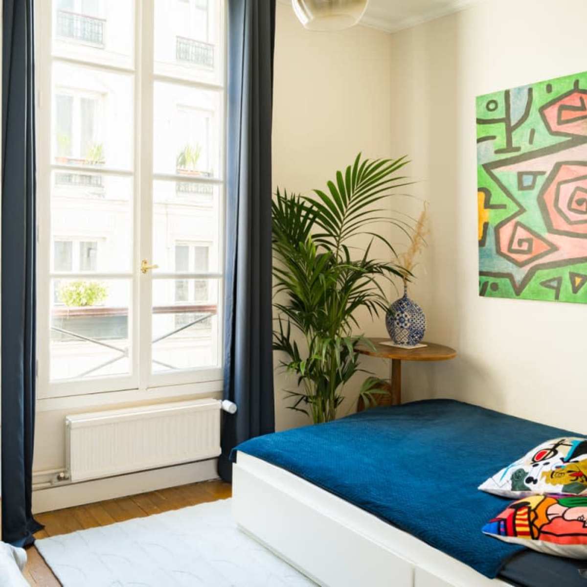 A bedroom with a blue bedspread, a potted plant, and a colorful abstract painting on the wall, illuminated by natural light from a large window.