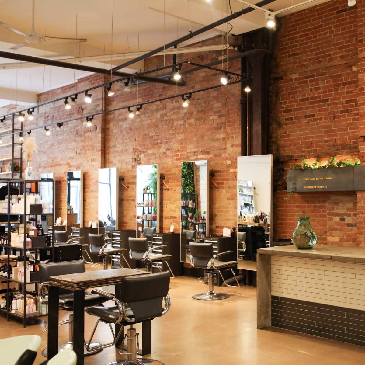 The interior of a modern hair salon with brick walls, styling stations, and a reception area featuring decorative shelves and a computer.