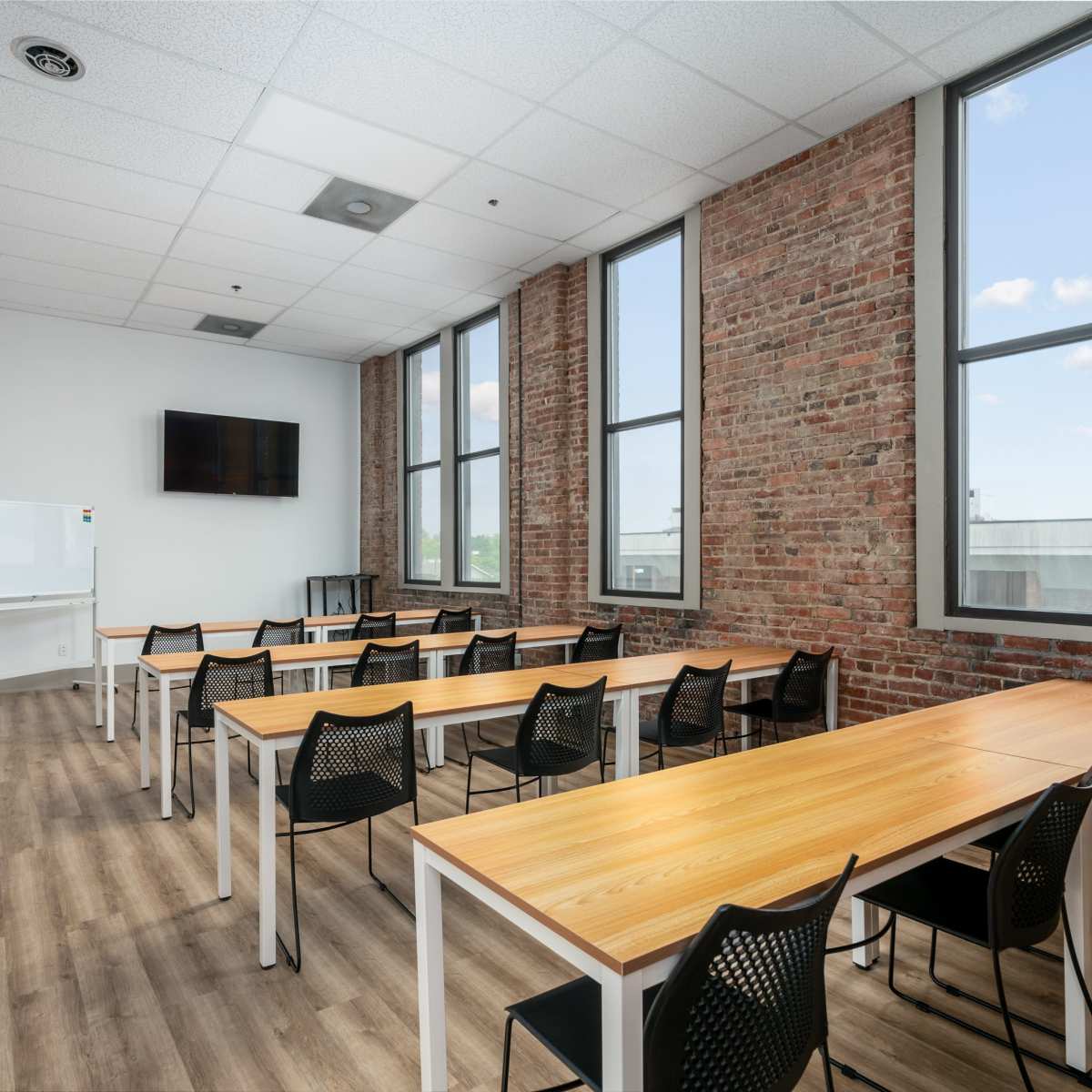 A modern classroom with wooden tables and black chairs, large windows, and exposed brick walls.