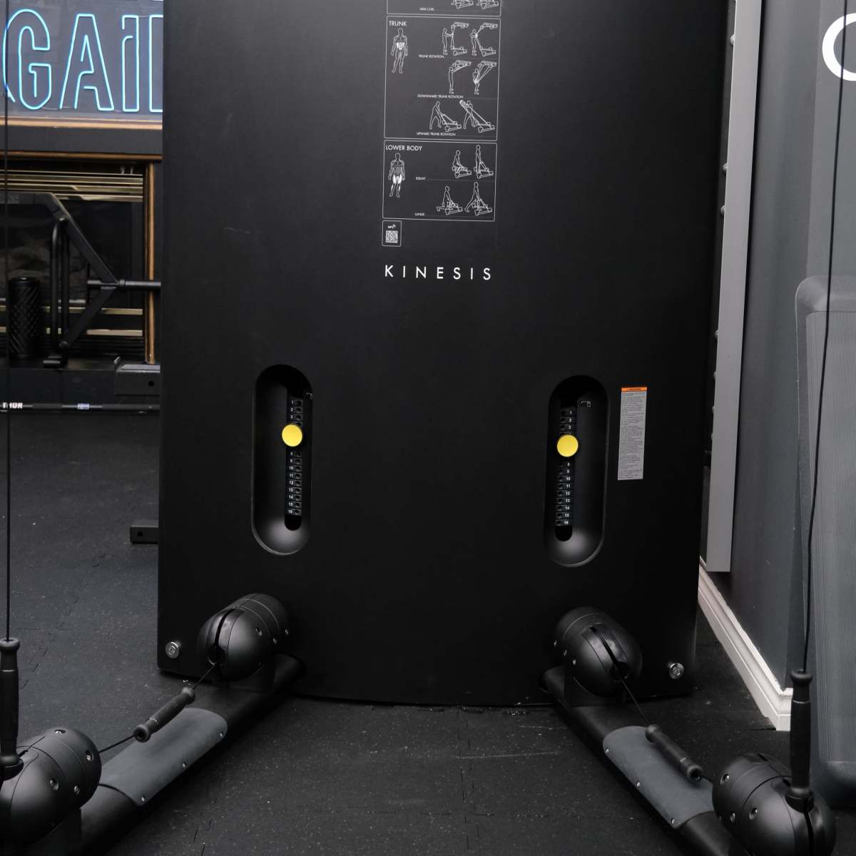A black Kinesis gym machine featuring multiple pulleys and instructional diagrams for exercises.