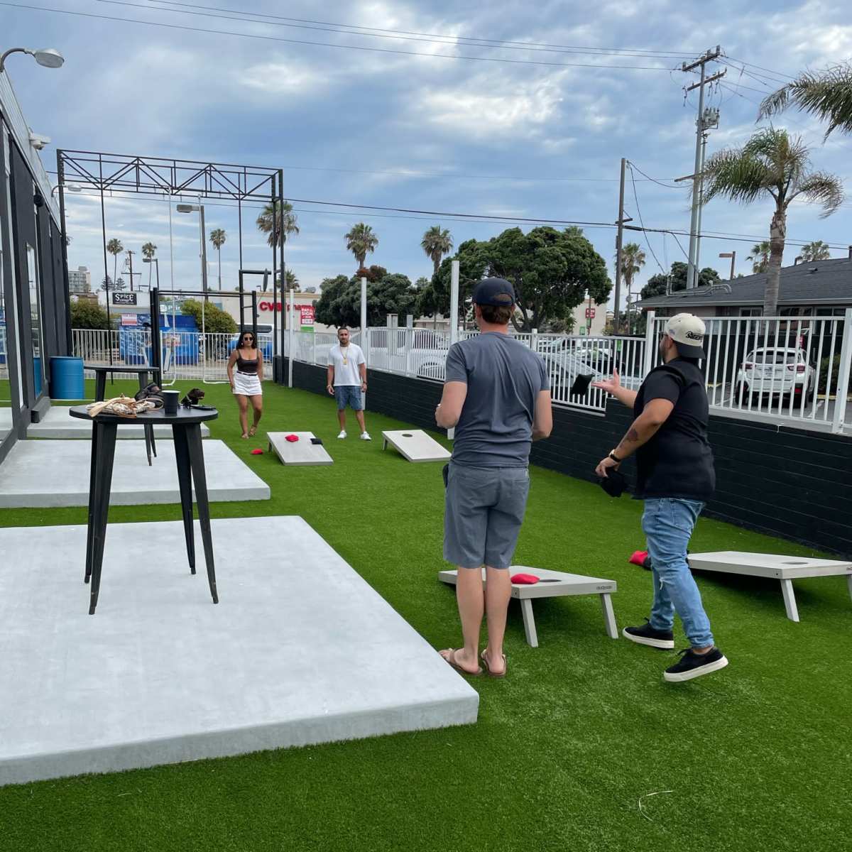Four people play cornhole on a grassy outdoor area with palm trees in the background.