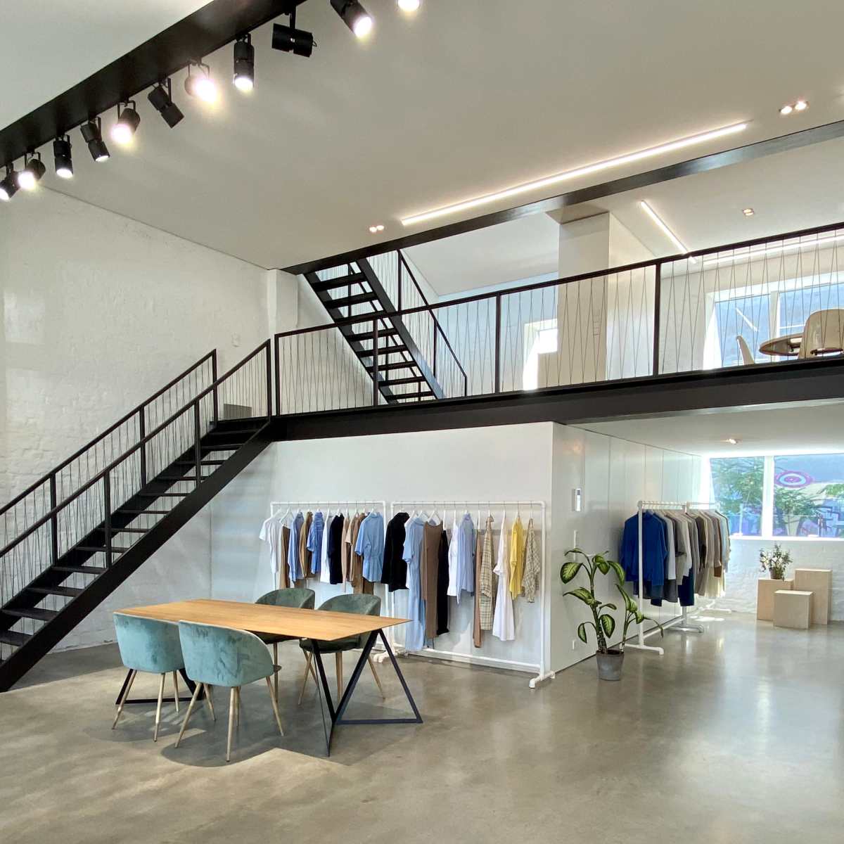 A modern clothing store features a wooden table with blue chairs, hanging garments on racks, and a staircase leading to an upper level.