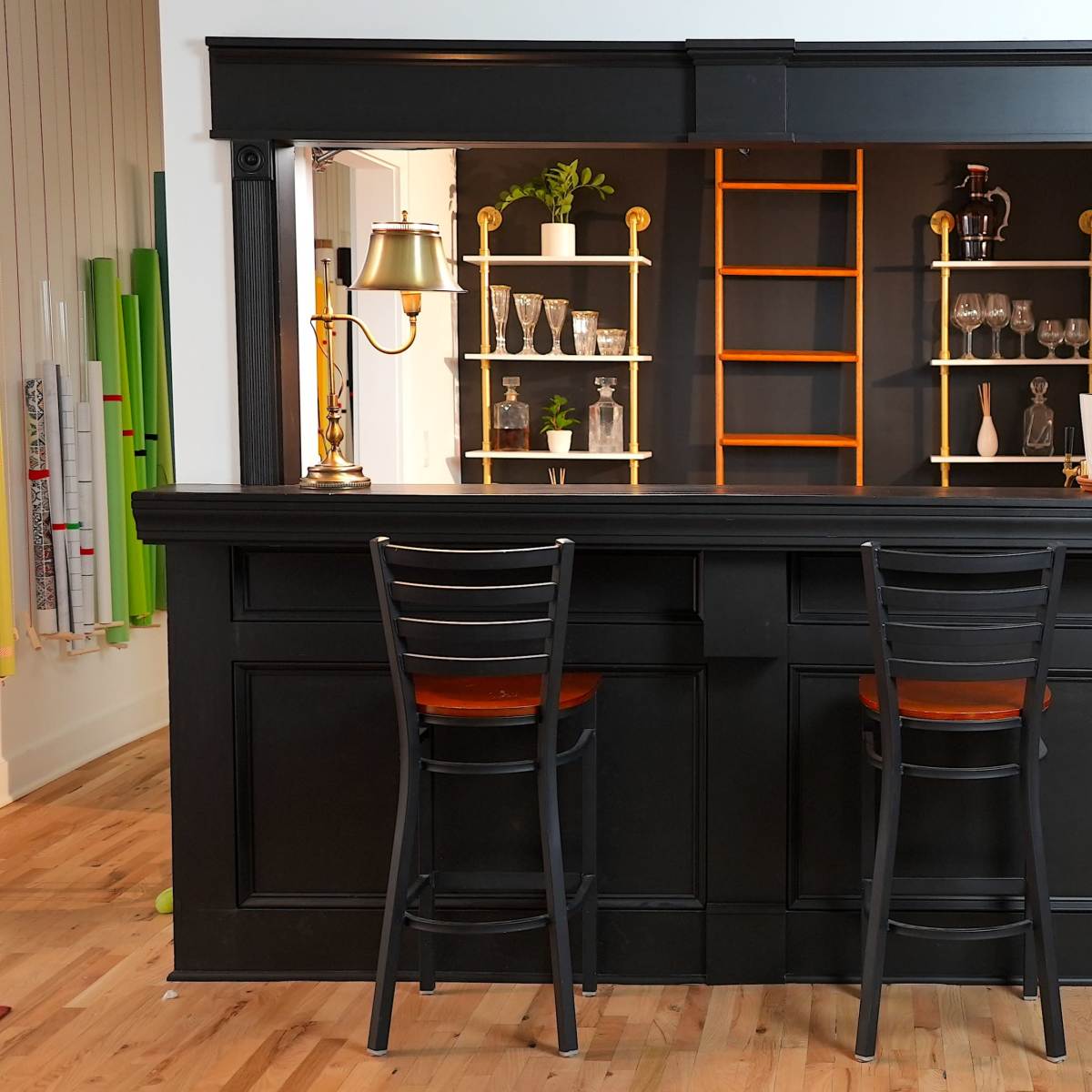 A black bar with two high stools in front, set against a wall featuring glass shelves holding various glassware, alongside a wooden door.