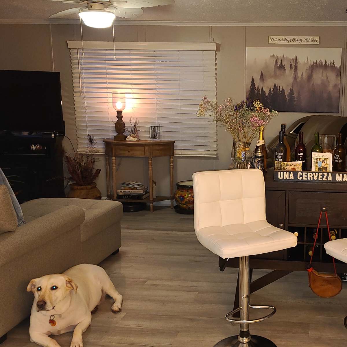 A cozy living space featuring a beige couch, two white bar stools, a bar cart stocked with various bottles, and a yellow dog lying on the floor.