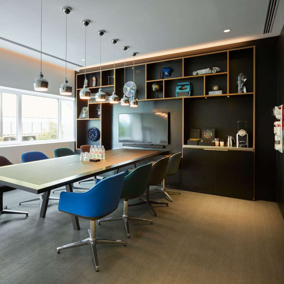 A modern conference room with a long table, assorted colorful chairs, a wall-mounted screen, and shelves displaying various items.