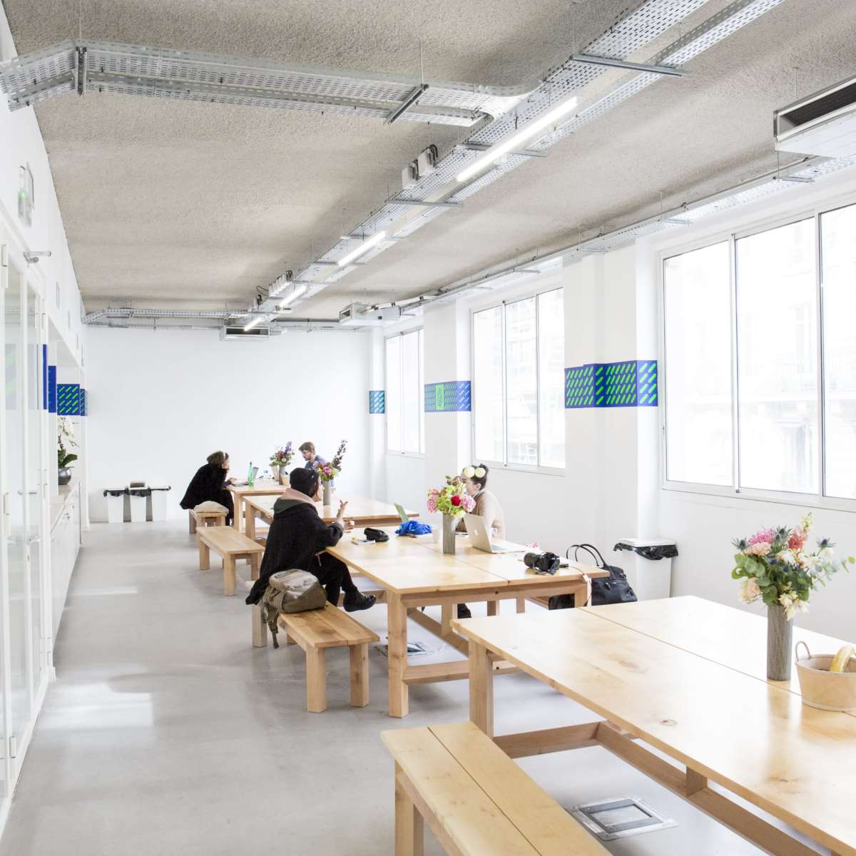 A brightly lit co-working space features long wooden tables with people working quietly and flowers decorating the center.