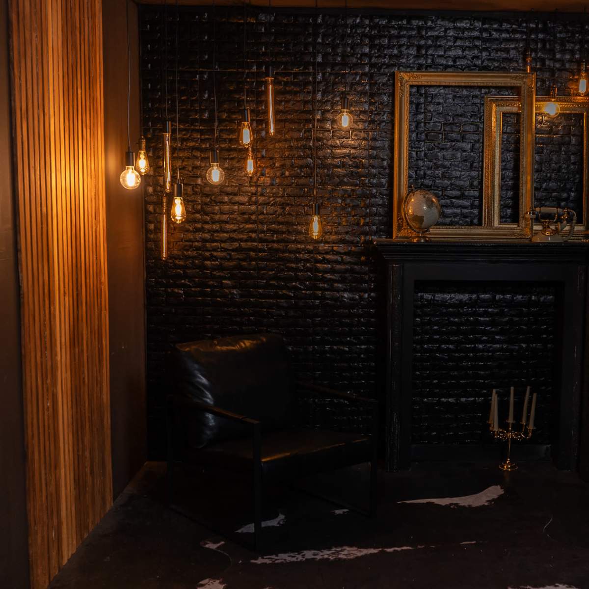 A dimly lit corner of a room featuring a black brick wall, a black armchair, hanging light bulbs, a decorative mirror, and a fireplace with a candle holder.